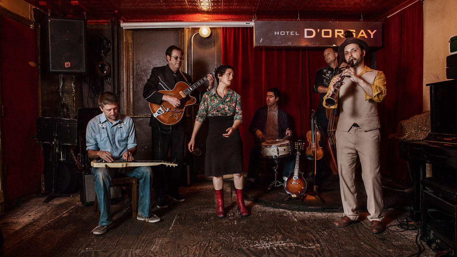 Six musicians in 1950s inspired outfits against a red velvet backdrop and a sign for Hotel D'Orsay.