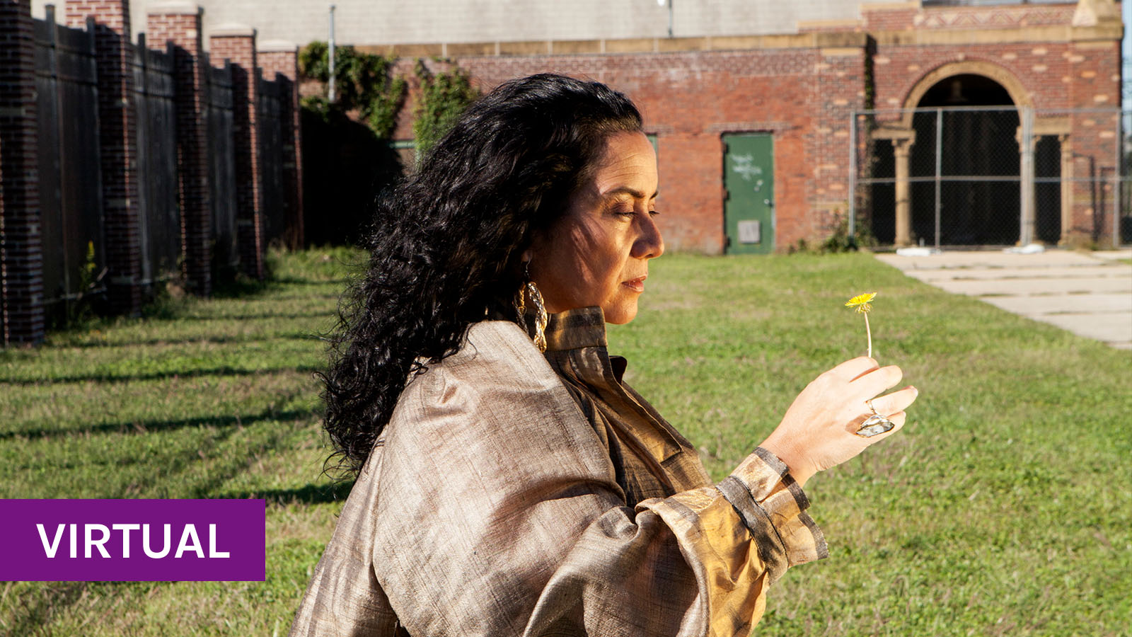 Claudia is standing on grass with a brick building behind her while holding a dandelion. She has  black curly hair and is wearing a patterned dress.