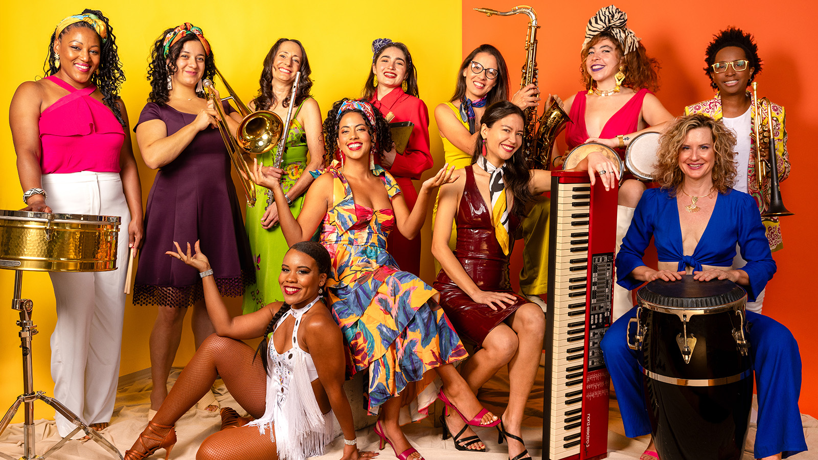 A group of 11 women pose with their instruments in colorful clothes infront of a yellow and orange background