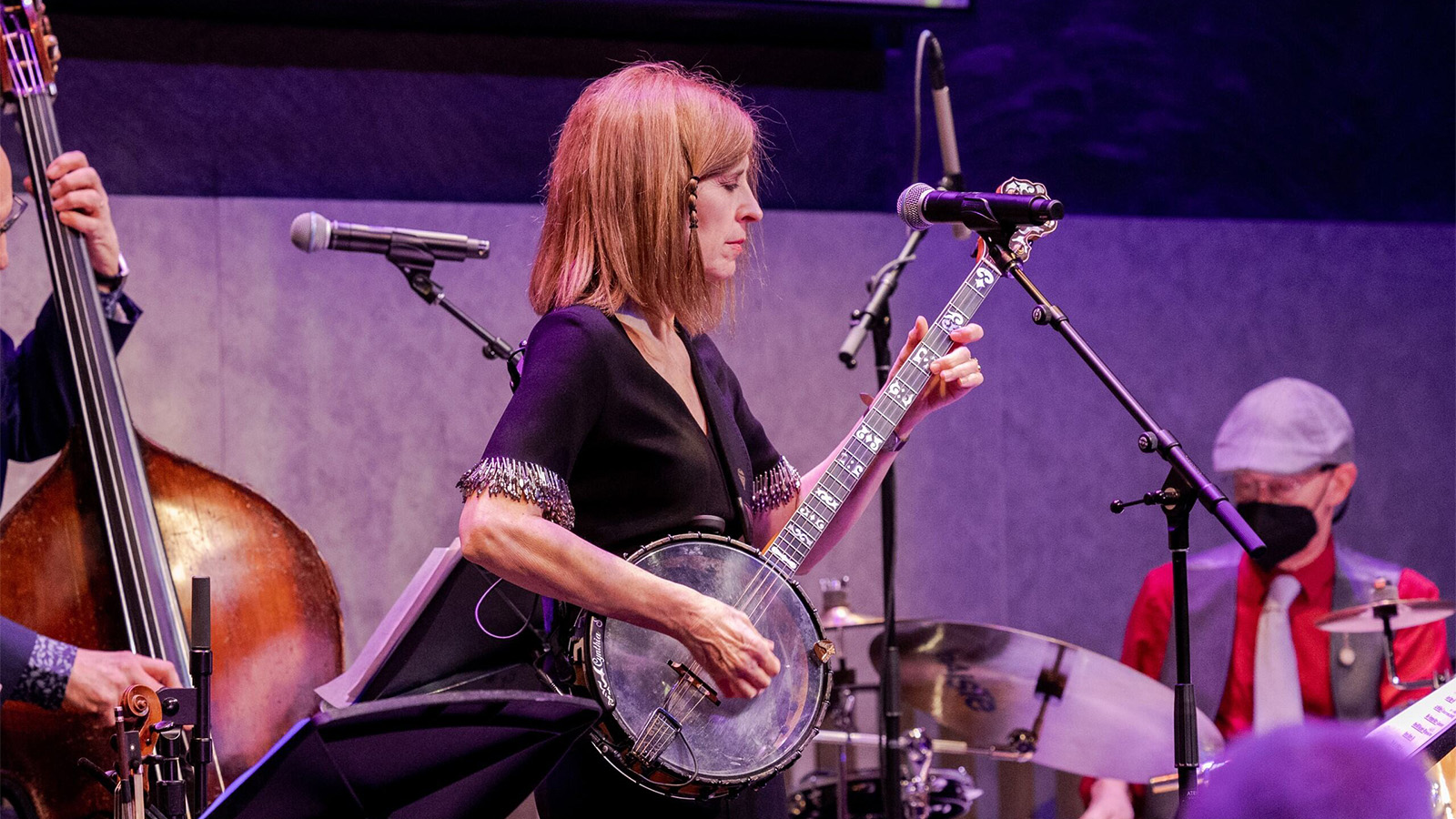 Cynthia Sayer stands onstage in front of a microphone looking down while playing a four-string banjo with a transparent drum. Behind her, members of Joyride Band play drums and upright bass.