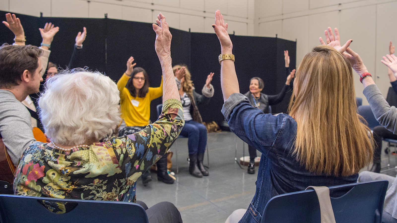 A dozen people, most older adults, sitting in a circle raise their arms in the air. One person holds a guitar.