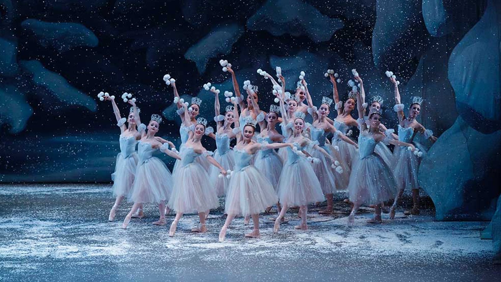 Sixteen ballerinas clustered on a snowy stage with a winter backdrop, wearing light bluish-white tutus hold clusters of white balls, raising arms upwards. 