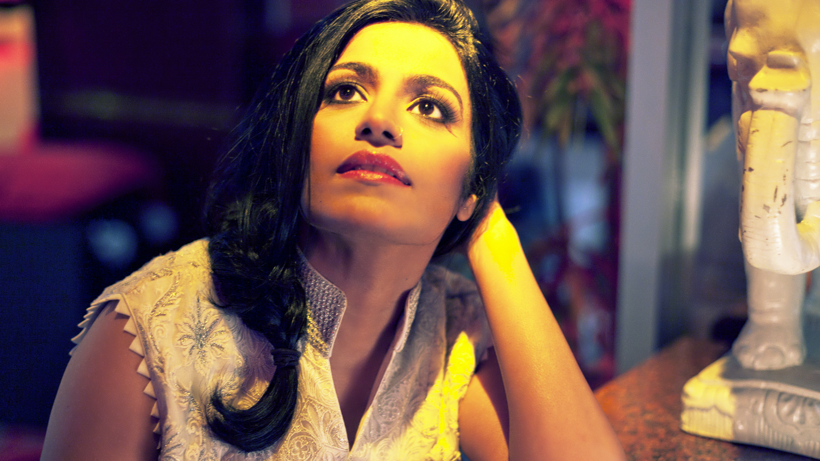 Close up of Falu wearing a white textured blouse and gazing upward. She is leaning with one arm on a table next to a white elephant statue.