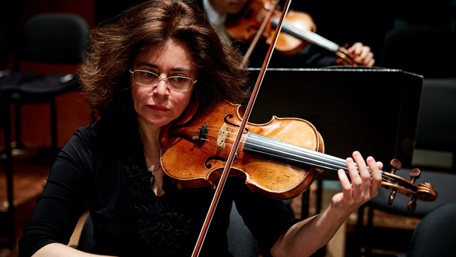Woman with short brown hair and glasses seated playing violin among other orchestra members.