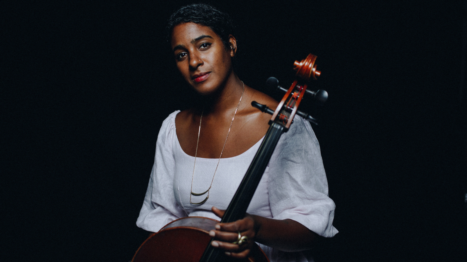Leyla McCalla looks directly into the camera with a soft smile, wearing a bright white top with wide sleeves and a thin silver necklace and holding the neck of a cello. Leyla glows against a dark black background.