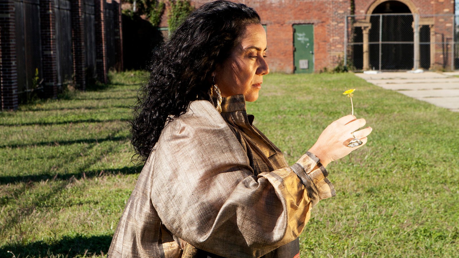 Claudia is standing on grass with a brick building behind her while holding a dandelion. She has  black curly hair and is wearing a patterned dress.
