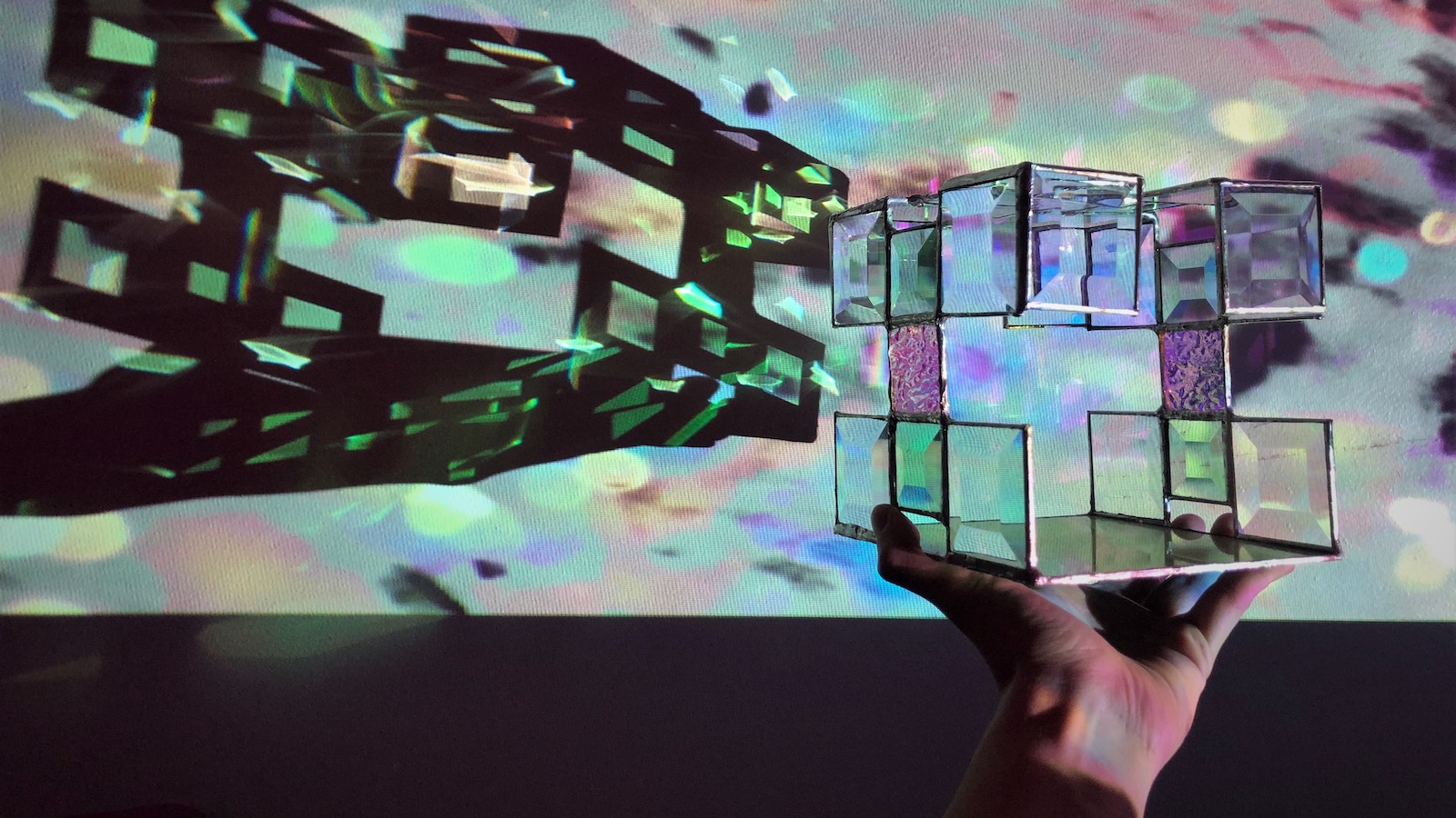 In a dark room, a hand holds a cube-like tesseract with square cutouts that refract multi-colored lights. The tesseract is held in front of a projector, casting a shadow of the cube onto a screen with twinkling circles of lights around it.