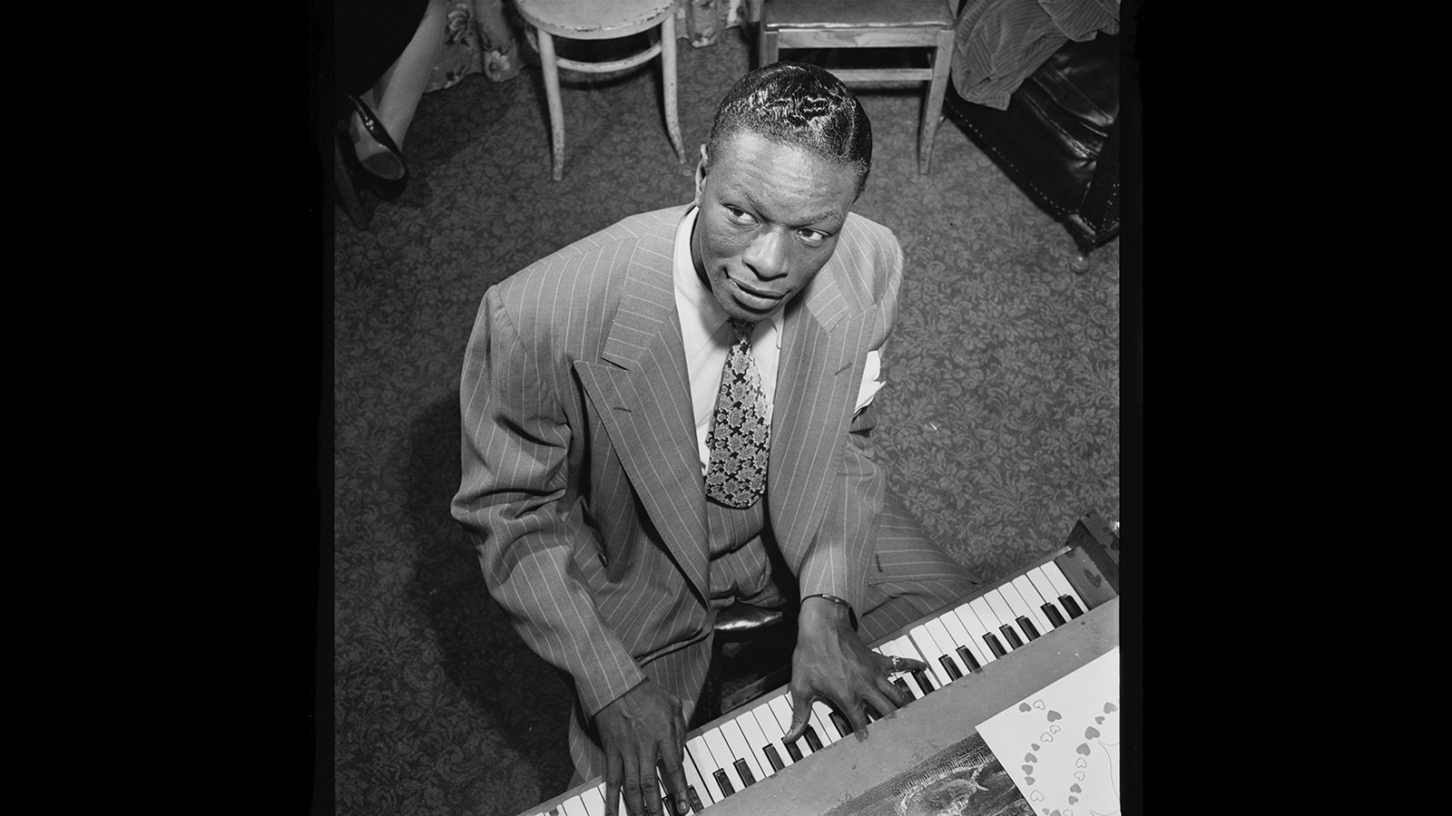 A black and white photo of Nat King Cole playing piano, while wearing a suit and tie.