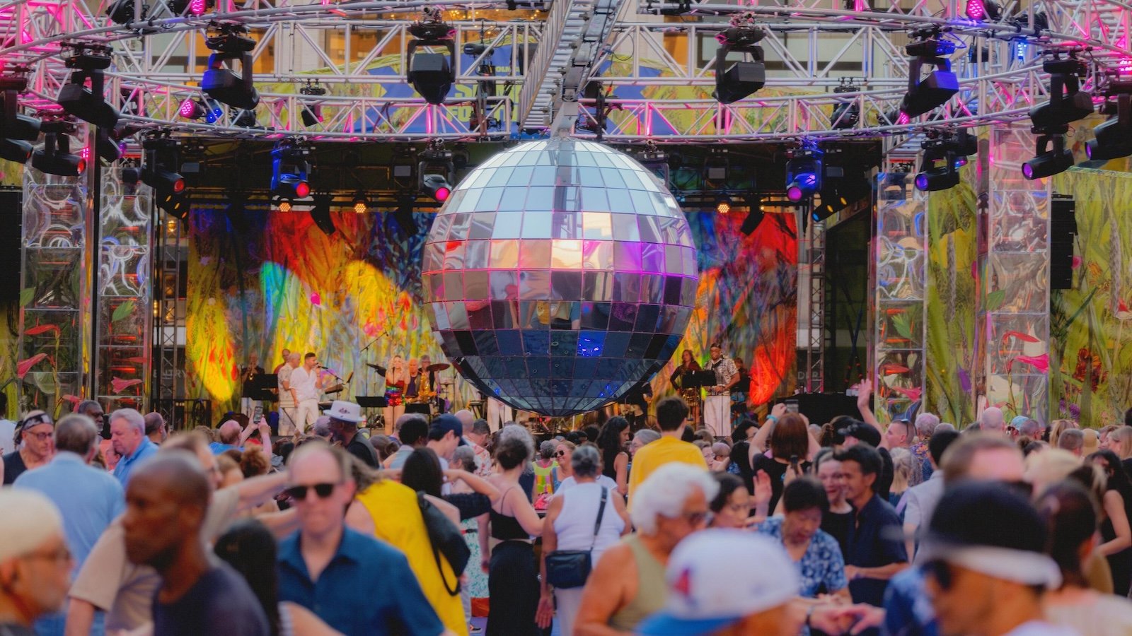 A crowd of people watch a performance onstage on Lincoln Center’s outdoor Dance Floor under a large disco ball lit pink. 