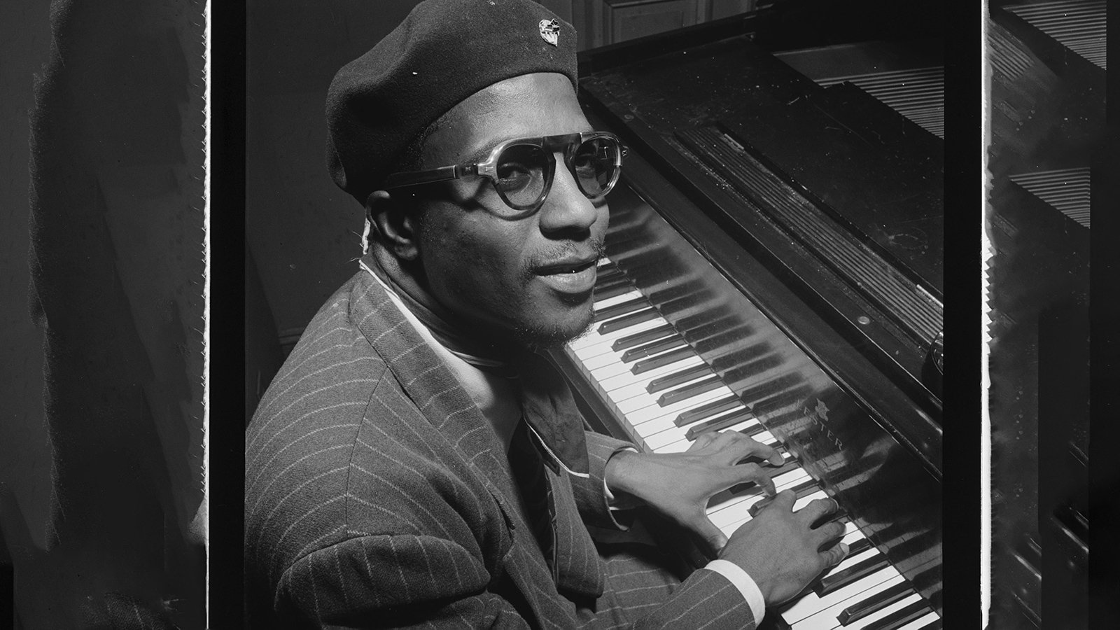A black and white photo of Thelonious Monk wearing a beret and lightly tinted sunglasses, sitting at a piano