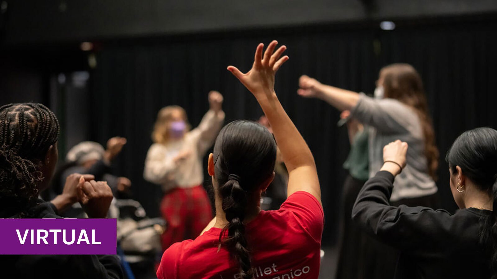 A person wearing a red Ballet Hispanico tshirt raises their hand watching two people moving in front of them.
