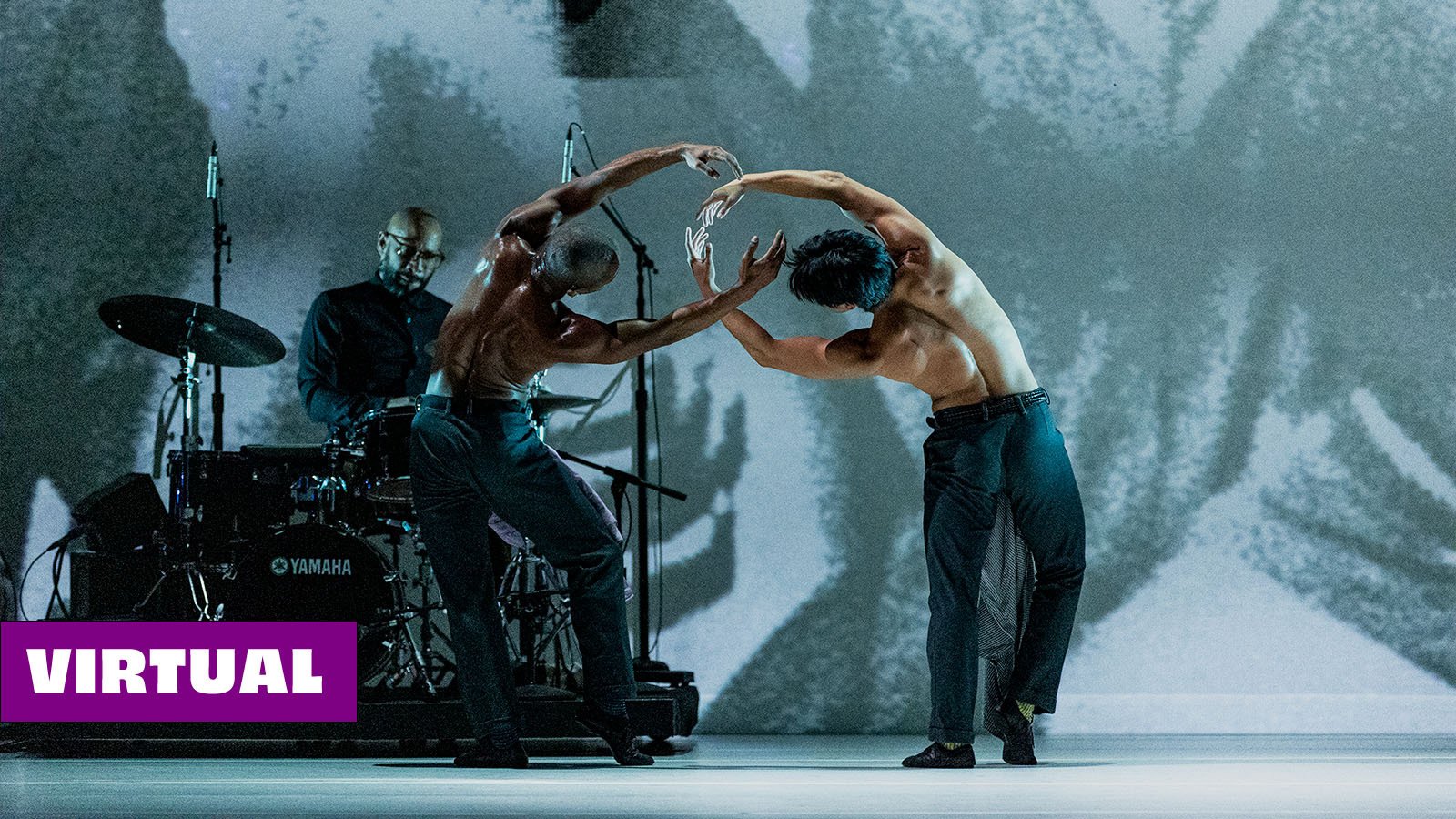 Two dancers perform onstage in front of a grey and white backdrop and a drummer playing a drum set. Shirtless with dark pants, the dancers stand with their backs to the audience leaning sharply towards the other. Their arms are held rounded above heads, interlocking between their bodies. 