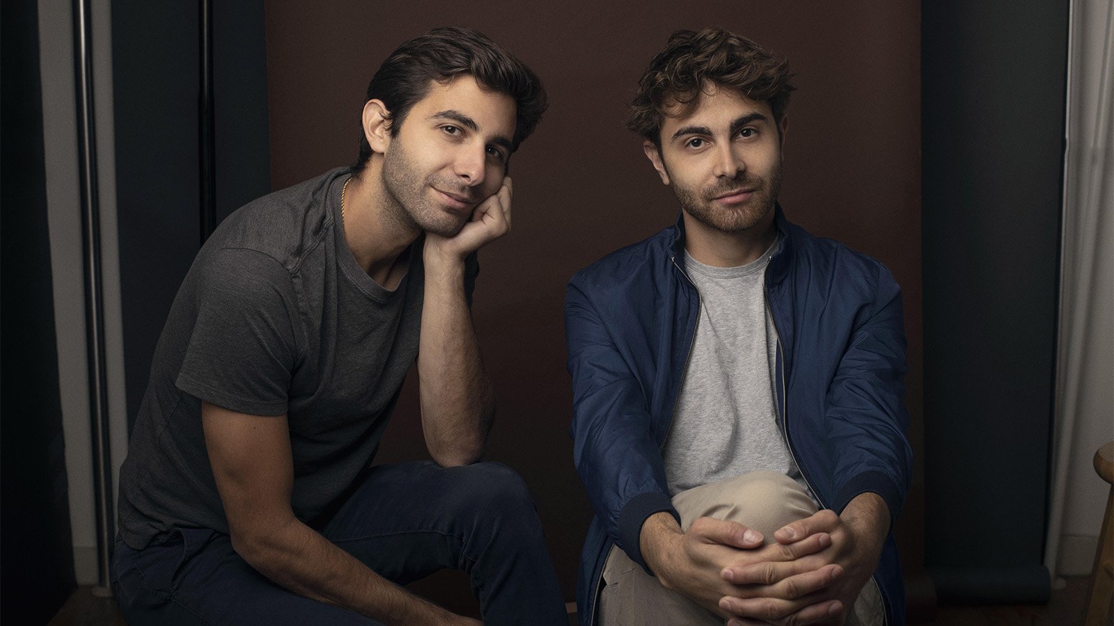 Patrick (left) and Daniel (right) Lazour sit in front of a brown background wearing street clothes in muted colors. Patrick has his head on his left hand, looking at the camera with a pensive expression. Daniel looks stoically at the camera with his legs crossed and hands clasped around his right knee