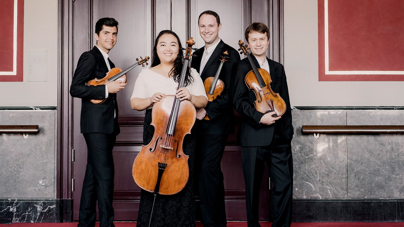 The members of the Callidore String Quartet stand in a hallway in front of a large wooden door wearing formal black and white clothes. From left to right, Ryan Meehan stands sideways, smiling at the camera and holding his violin, Estelle Choi smiles at the camera holding her cello in front of her, Jeffrey Myers stands behind Estelle, smiling at the camera with his violin tucked under his left arm; and  Jeremy Berry stands back-to-back with Jeffrey Myers, resting his viola on his right hand and right shoulder. 