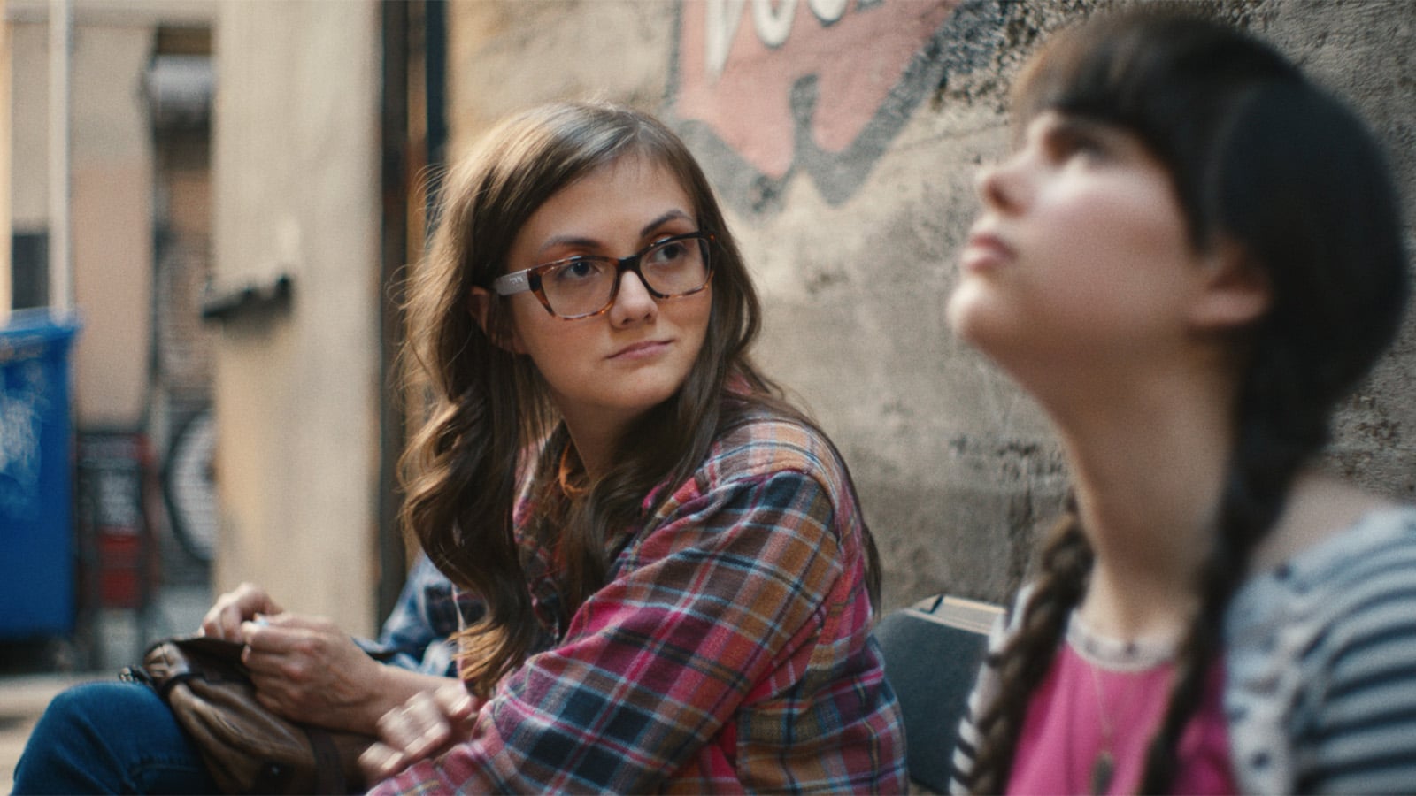 Two young women sit on a bench in an alley. One wears glasses and a plaid shirt, looking thoughtfully at the other, who has long braids and a pink shirt while gazing upwards. A blue dumpster and graffiti are visible in the background.